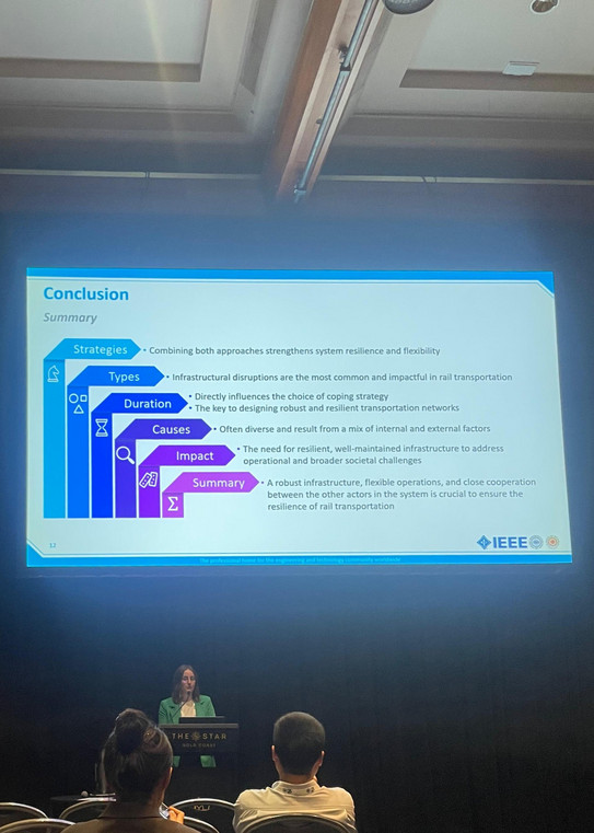 Franziska Rosenthal steht auf einer Bühne hinter einem Pult. Oben über ihr sieht man eine Folie. Vor der Bühne stehen Stühle auf denen Zuhörer sitzen.