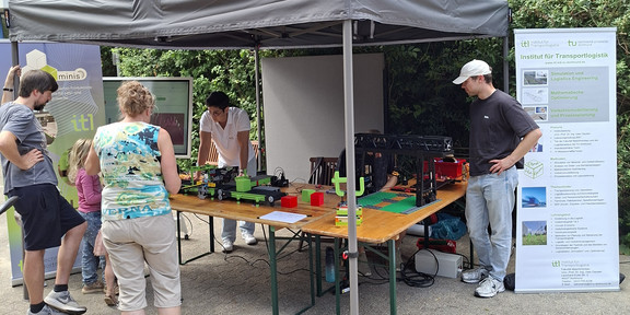 Mehrere Menschen stehen vor einem Pavillon unter dem Tische aufgebaut wurden, auf denen Ausstellungsstücke stehen