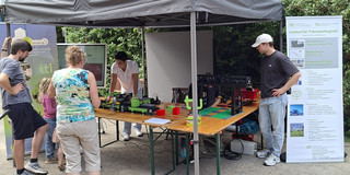 Mehrere Menschen stehen vor einem Pavillon unter dem Tische aufgebaut wurden, auf denen Ausstellungsstücke stehen