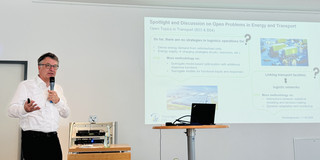 Professor Clausen hält ein Mikrofon. Hinter ihm ist das Bild einer Präsentation zu sehen.
