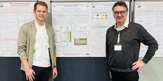 Professor Clausen und Herr Mowe stehen vor einer Pinnwand mit einem Poster