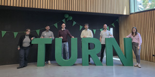 Personen stehen auf einer Bühne vor einer Holzwand hinter eine Plastik die das Wort TURN bildet.