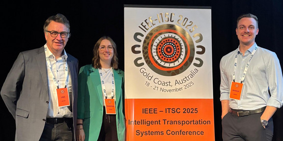 Professor Clausen, Franziska Rosenthal und Marius Dellbrügge stehen vor einem Werbeposter der Konferenz