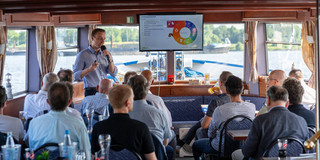 Dr. Marius Dellbrügge steht vor einer Menschengruppe die sitzt in einem Schiff und hält einen Vortrag.