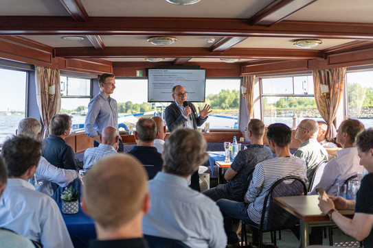 Dr. Dellbrügge steht neben einem Herrn der eine Rede hält auf einem Schiff. Eine Menschengruppe schaut zu.