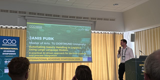 Herr Janis Purk steht auf einer Bühne und hält ein Mikrofon. Neben ihm ist eine Leinwand mit einer Präsentation zu sehen.