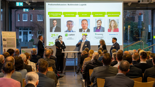 Teilnehmer einer Podiumsdiskussion stehen vor einer Leinwand und diskutieren. Im Vordergrund sitzt Publikum und schaut zu.