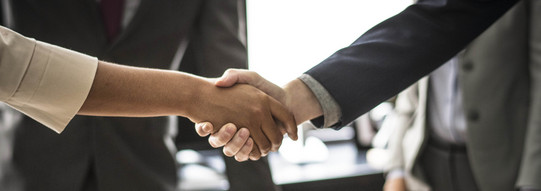 Zwei Personen geben sich die Hand. Das Händeschütteln ist im Detail abgebildet, im Hintergrund stehen zwei Menschen im Anzug.
